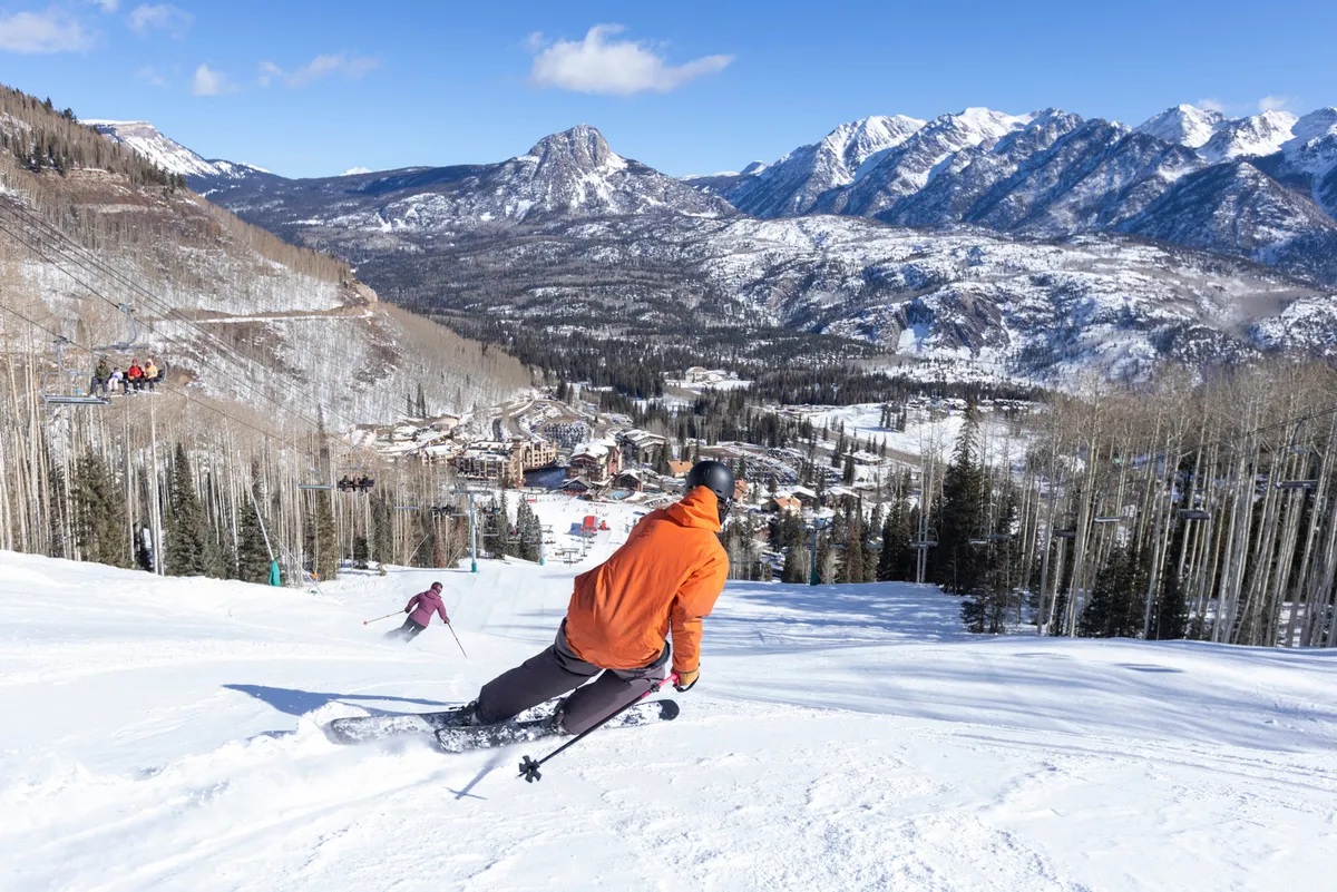 Dynamic action shot of two skiers—one in an orange jacket, the other in purple—carving down a snowy mountain, with the resort village and rugged peaks visible below. The scene pulses with energy, capturing the thrill of skiing and the panoramic beauty of the surroundings.