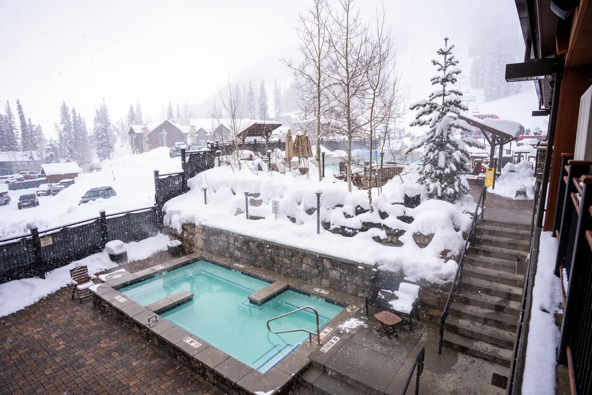 A tranquil heated outdoor pool set against a snow-covered landscape, surrounded by trees. A staircase leads down to the pool area, where guests relax in the warm water while taking in the winter scenery. The atmosphere is serene and inviting, emphasizing relaxation and a connection to the natural mountain surroundings.