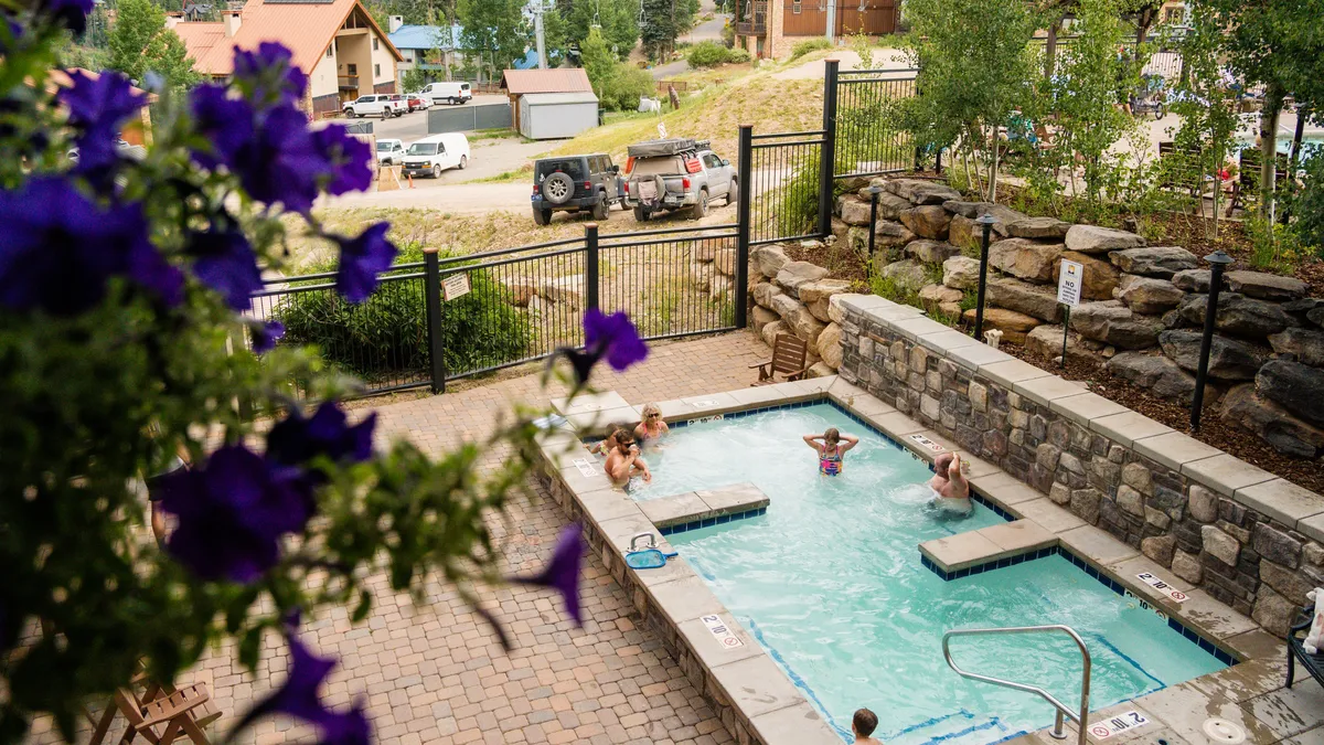 A vibrant outdoor scene featuring a heated swimming pool surrounded by blooming flowers, with children laughing and playing in the water. Adults relax on sun loungers nearby, while a rock wall provides a scenic backdrop, creating a lively and inviting atmosphere perfect for families.