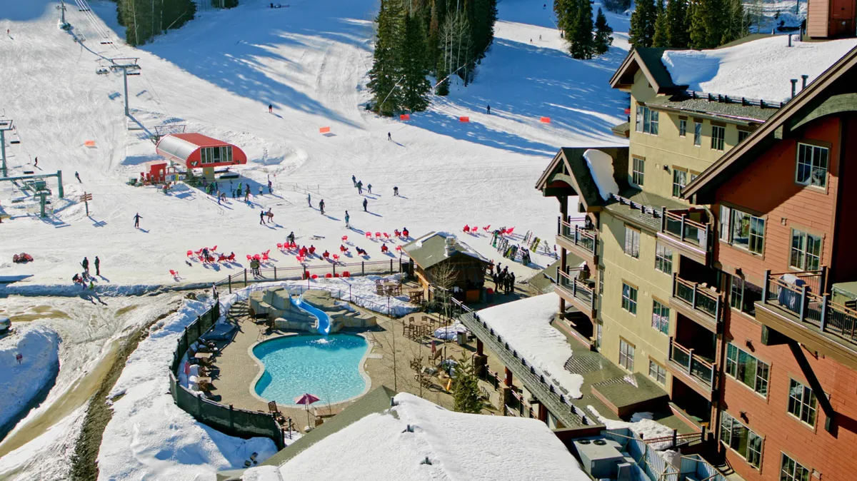 A lively exterior view featuring a swimming pool positioned beside ski slopes and lifts, with the lodge's architecture in the background. The scene conveys both excitement from mountain activities and the relaxation of poolside moments, blending adventure and leisure.