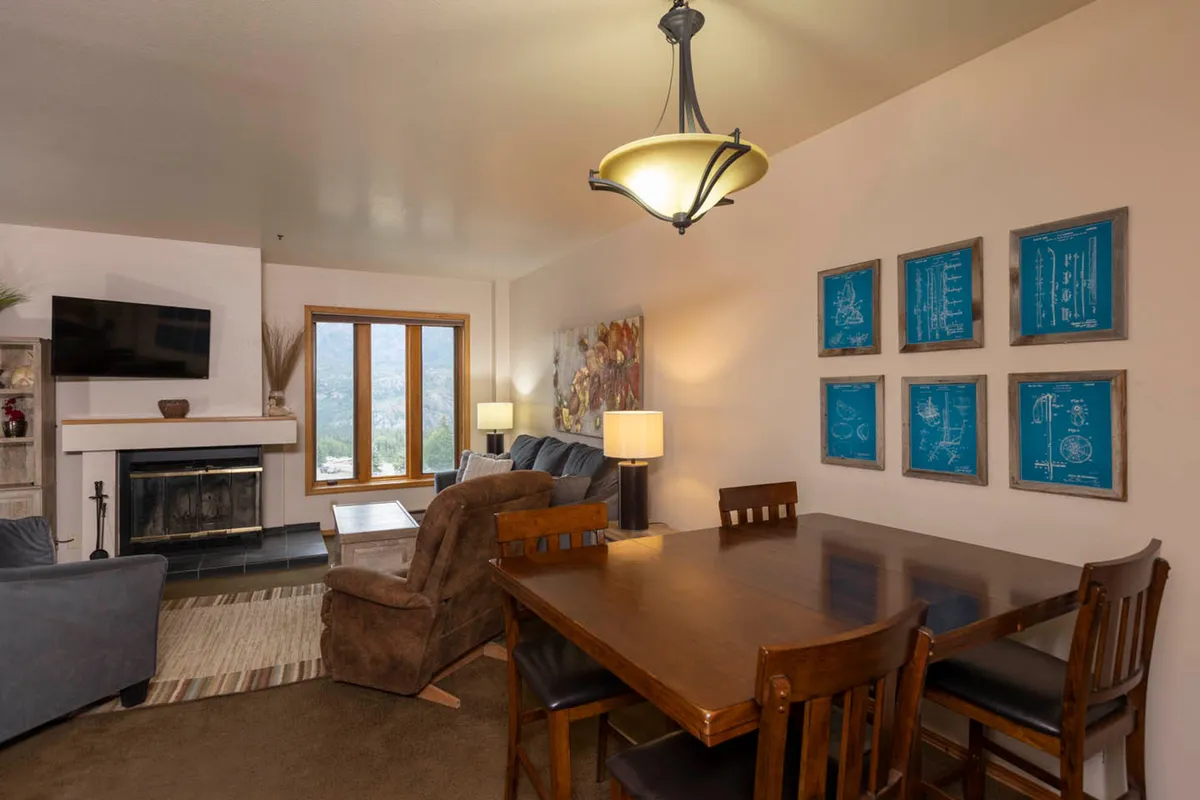 This scene focuses on an open-concept space combining a dining area and a living area with a fireplace. The dining table sits near the fire, with soft lighting and mountain views through the windows, creating a seamless setting for shared meals and lively conversation.