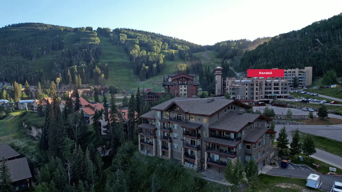 A wide exterior shot showcasing the mountain resort buildings framed by a dramatic mountain backdrop and lush tree line, with visible parking area and welcoming lodge atmosphere. The setting hints at both adventure and relaxation, with the mountains beckoning just beyond the resort.