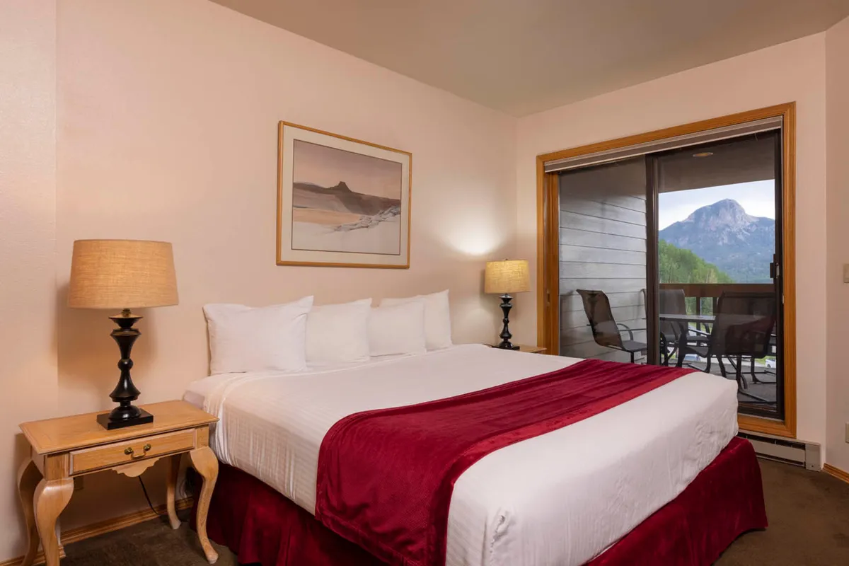 A cozy bedroom with a bed dressed in white linens and accented by a red runner, a table lamp, and a sliding door revealing a breathtaking mountain view. Warm lighting gives the room an inviting glow, perfect for a peaceful retreat.