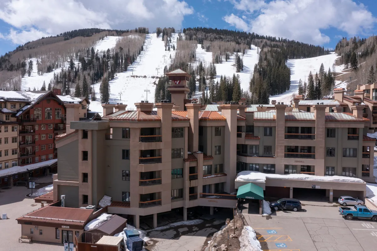 This exterior image captures the condo building, its adjacent parking area, and the breathtaking snow-covered mountain backdrop. The parking area is clearly visible, illustrating the convenience of on-site, presumably garage, parking. The building itself suggests easy access to resort amenities and the surrounding natural beauty.