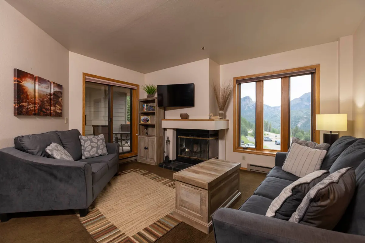 The image showcases a warm, inviting living space in a Durango condo, with a crackling wood-burning fireplace as the centerpiece. Plush sofas are arranged for cozy gatherings, and expansive windows frame stunning mountain views, basked in golden sunset light. The atmosphere is perfect for sipping a hot drink, unwinding after a day of alpine adventure, or sharing stories with loved ones.