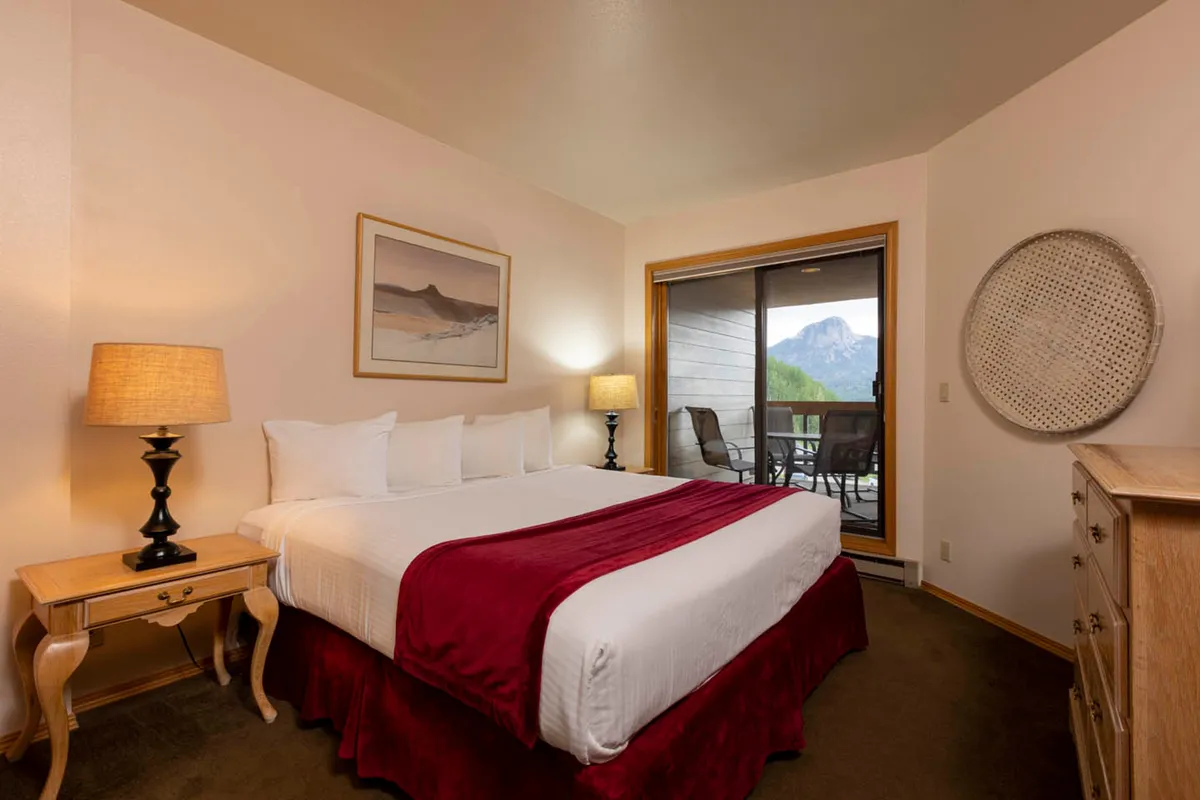 A serene bedroom featuring a queen-size bed with crisp white linens, a wooden dresser, and a nightstand with a lamp. A large window fills the space with natural light and offers a stunning mountain view, enhancing the calm and cozy ambiance.