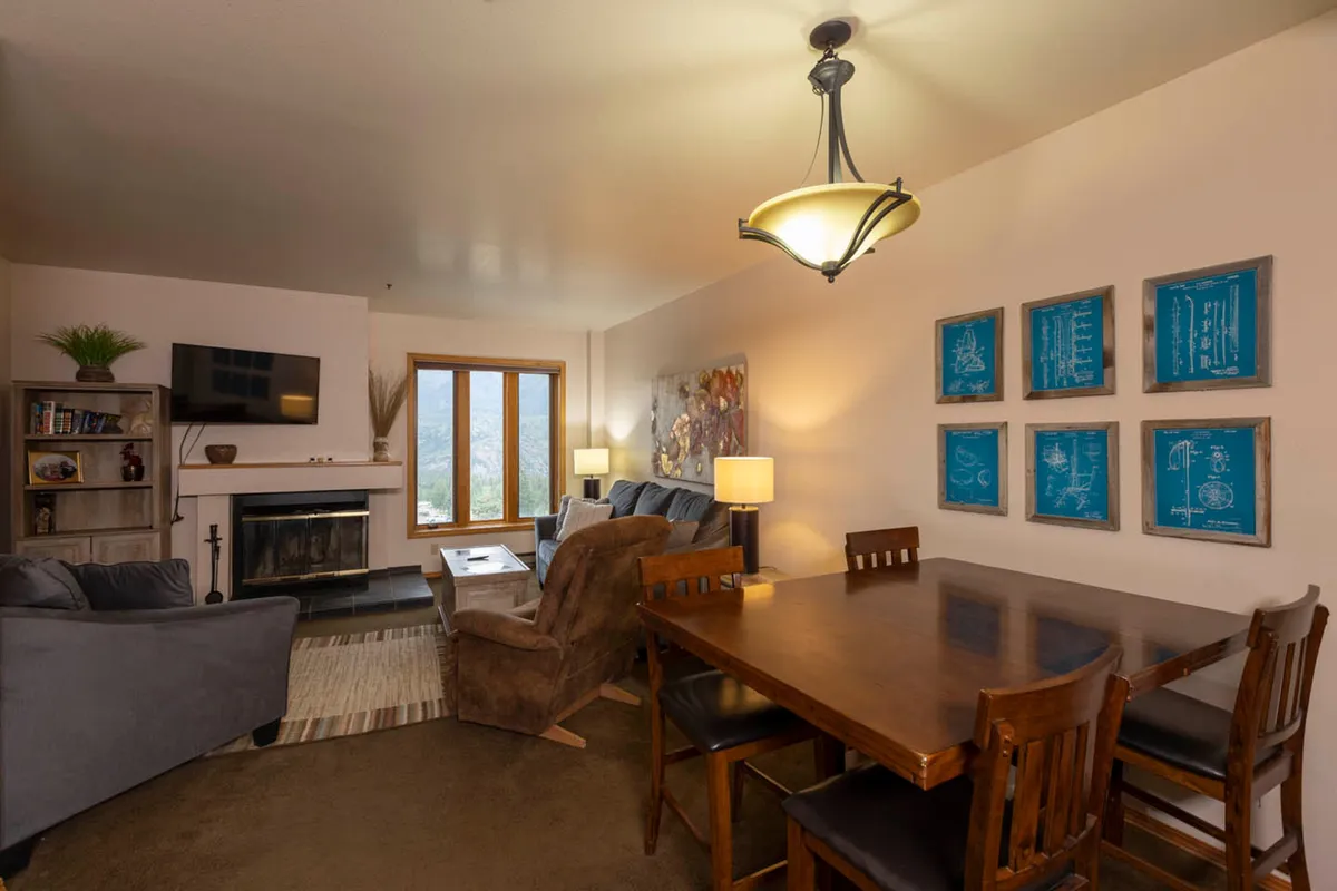 A warmly lit living area featuring a rustic wood-burning fireplace as the focal point. The space includes a cozy dining table set for two, inviting artwork on the walls, and large windows that hint at scenic views beyond. The overall mood is intimate and welcoming, perfect for relaxing after a day of outdoor adventures.