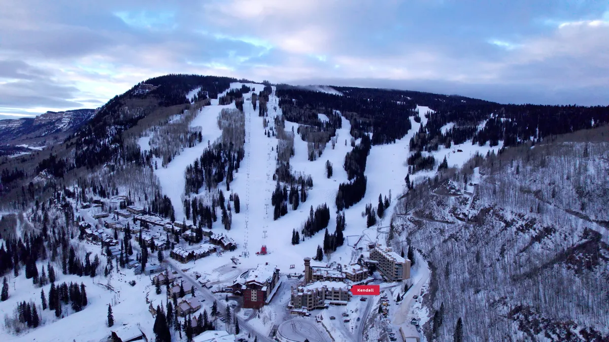 A panoramic view featuring snow-covered ski slopes winding down the mountain, surrounded by dramatic peaks and cozy lodging facilities nestled at the base. The scene is bathed in crisp mountain light, evoking the excitement of an alpine adventure and the allure of immediate slope access.